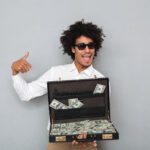 Image by drobotdean on Freepik. A black man with afro hair holding brief case full of money. He is wearing semi-formal clothes. Portrait of a happy young afro american man in sunglasses pointing finger at