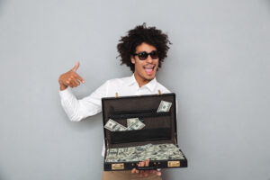 Image by drobotdean on Freepik. A black man with afro hair holding brief case full of money. He is wearing semi-formal clothes. Portrait of a happy young afro american man in sunglasses pointing finger at